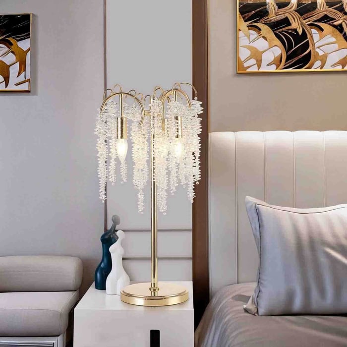 Gold multi-arm table lamp with cascading clear crystal strands on a bedside table beside a padded headboard and sofa.