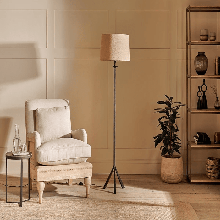 Neutral living room with a beige upholstered armchair, round side table with glass carafe, black metal tripod floor lamp with tan linen shade, potted plant, and open shelving against a paneled wall.