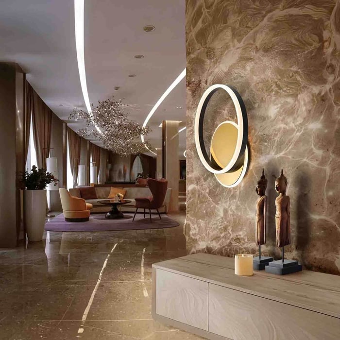 Modern luxury lobby with marble wall, glowing circular wall sconce, and a console table with decorative statues, leading into a softly lit seating area.
