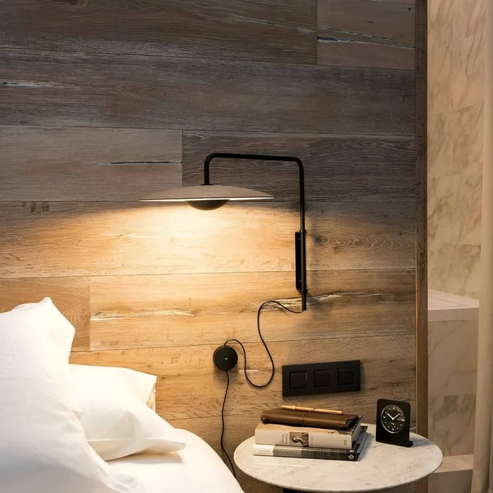 Black plug-in wall light with disc shade over a bedside table on a warm wood plank wall, white bedding and marble nightstand.