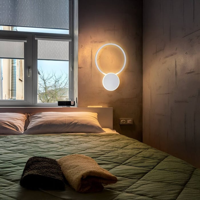 Minimalist circular LED wall fixture with halo effect, mounted beside a bed with green quilted bedding and folded towels.