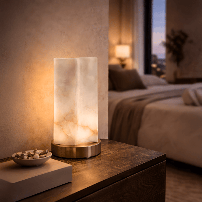 Cylindrical alabaster table lamp with natural veining on a wooden bedside table in a softly lit bedroom setting.