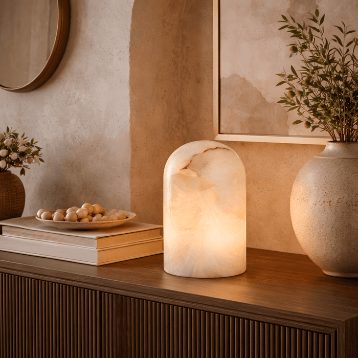 Sculptural arched alabaster table lamp glowing warmly on a wooden sideboard styled with books and decorative ceramics.