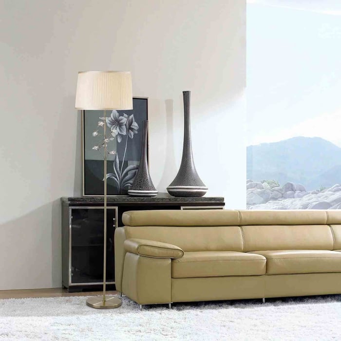 Slim brass floor lamp with pleated cream shade next to a beige leather sofa in a bright, modern lounge.