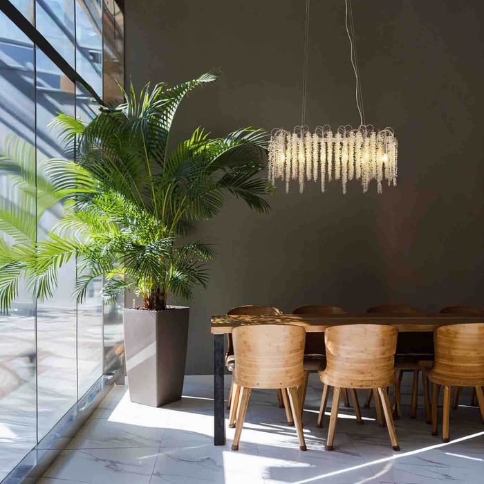 Crystal fringe linear chandelier glowing above a dining table beside a large potted plant and floor-to-ceiling windows.