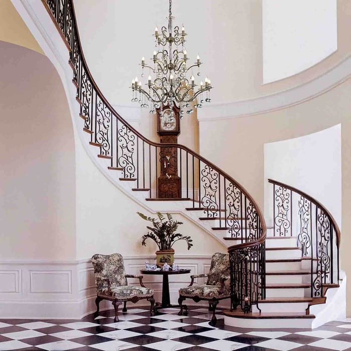 Large traditional chandelier illuminating a grand foyer with a sweeping curved staircase, ornate iron balustrade, and black-and-white tiled floor.