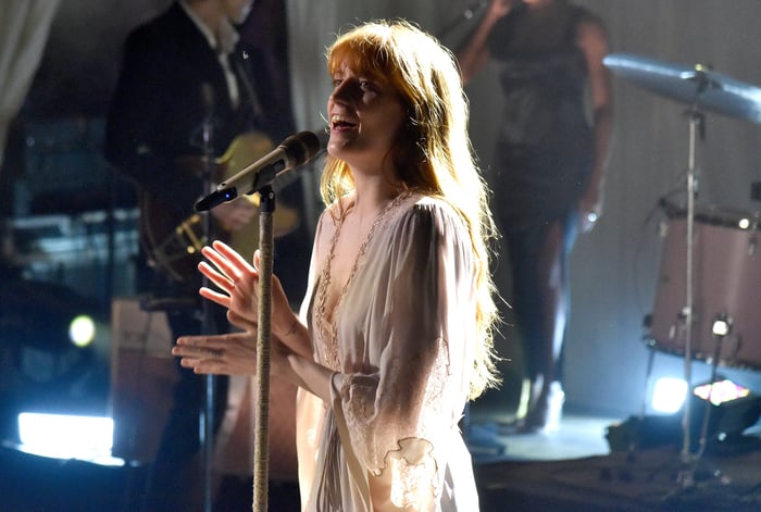 Florence Welch in red dress digging a hole in a desolate landscape