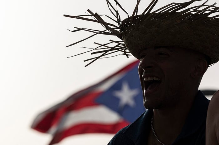 A man wears a straw hat with the Puerto Rican flag in the background before the start of the first show of Puerto Rican singer Bad Bunny’s 30-date concert residency at the Coliseo de Puerto Rico in San Juan, on July 11, 2025. (Credit: Ricardo ARDUENGO / AFP)
