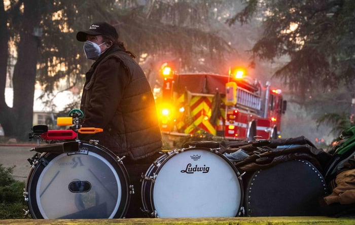 A man watches his home burn while sitting with a drum set and other belongings on Santa Rosa Avenue in Altadena on January 8, 2025.