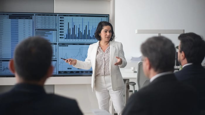  woman in a white suit is presenting data on a large screen, which includes financial charts, graphs, and tables. The audience, dressed in business attire, is attentively listening.