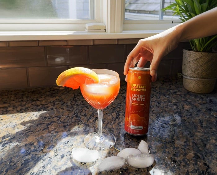 Pink Blood Orange Sunset mocktail in a cut-glass coupe with a blood orange wedge, beside a hand holding a can of Upstate Elevator Lychee Blood Orange Uplift Spritz on a sunny kitchen counter