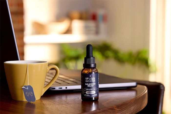 Upstate Elevator Balanced Relief hemp extract bottle beside a yellow mug and laptop.