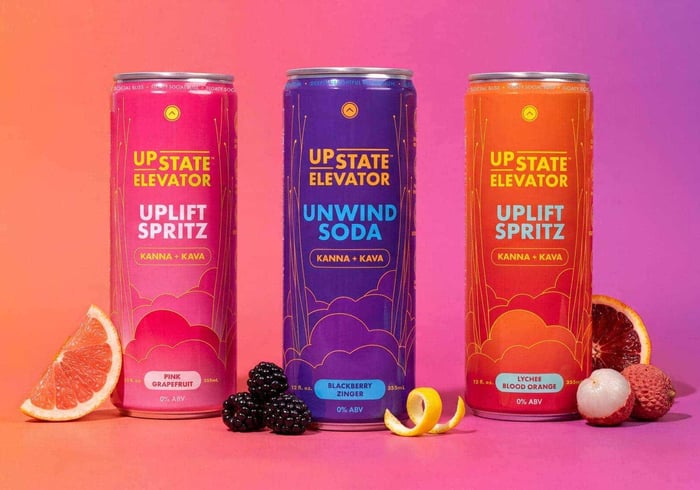 Pink Grapefruit Uplift Spritz, Blackberry Zinger Unwind Soda and Lychee Blood Orange Uplift Spritz cans on a pink backdrop with grapefruit, blackberries, lemon peel, lychee and blood orange