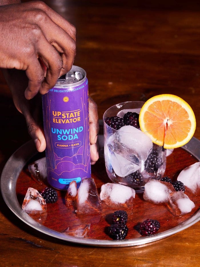 Hand reaching for a can of Upstate Elevator Blackberry Zinger Unwind Soda on a tray with ice, blackberries and an orange slice