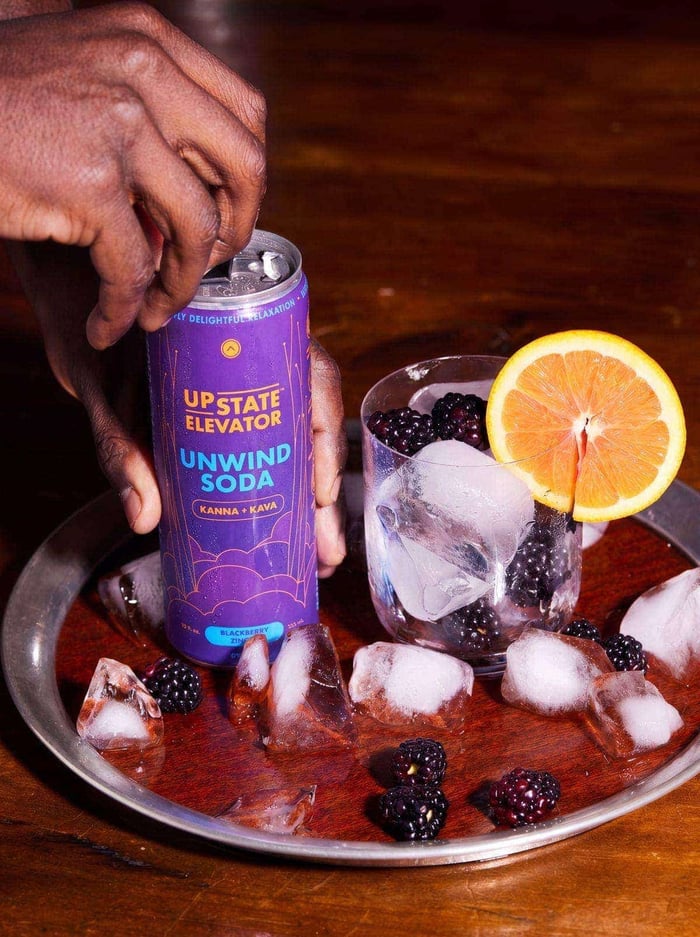 Hand lifting a can of Upstate Elevator Blackberry Zinger Unwind Soda from a tray with a glass of ice, blackberries and an orange slice