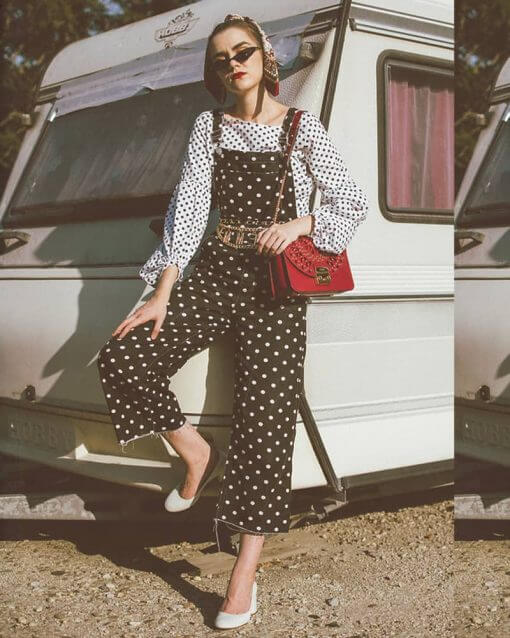 Woman wears polkadot print shirt under polkadot print overalls with bright red bag and many accessories.