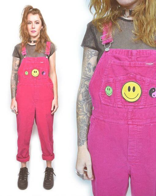Woman wears neon pink overalls with smiley face and yin yang patches over a black mesh top.