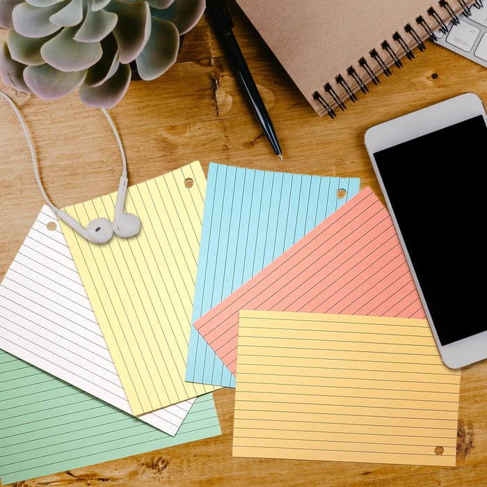 Colorful hole-punched 4x6 index cards spread out on a wooden desk with a smartphone, spiral notebook, pen, earbuds, and a succulent plant.
