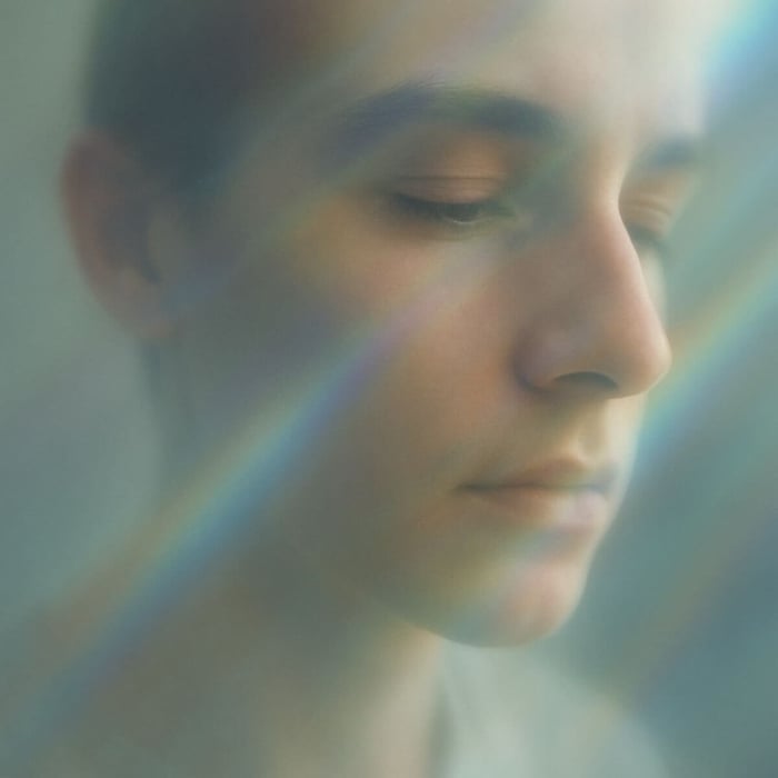 A serene human face seen through softly refracted glass, illuminated by arcs of violet, turquoise, and gold light. The person gazes calmly toward the luminous colors, conveying awareness, stillness, and receivership.