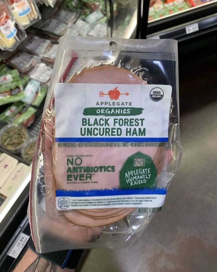 A hand holding a package of deli meat branded Applegate | Organics Black Forest Uncured Ham. 