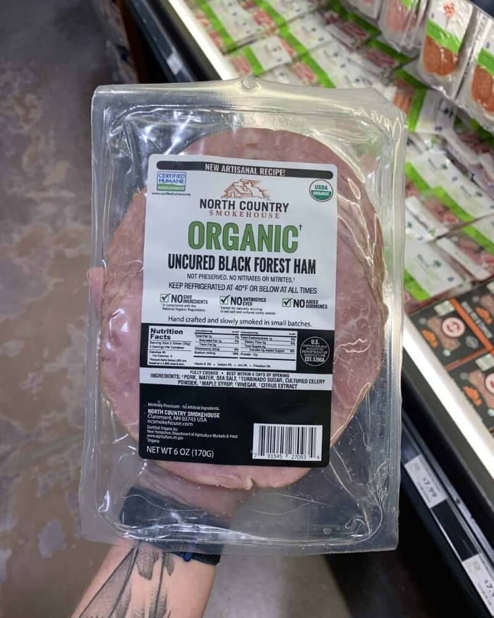 A hand holding a package of deli meat branded by Organic Uncured Black Forest Ham.