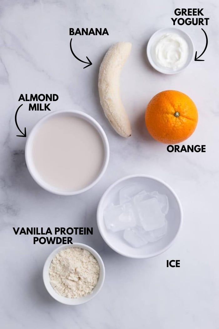 A photo with ingredients for orange protein shake laying flat against a white marbled surface.