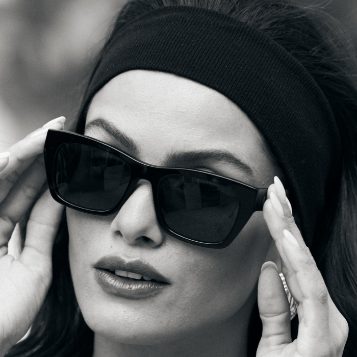 Close-up portrait of woman modeling oversized sunglasses 2025 with large black rectangular frames, hands positioned around face, wearing black beanie in monochrome photography