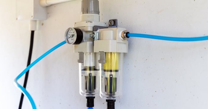 Compressed air filter regulator with pressure gauge and dual filter bowls connected to blue pneumatic tubing on a wall panel.