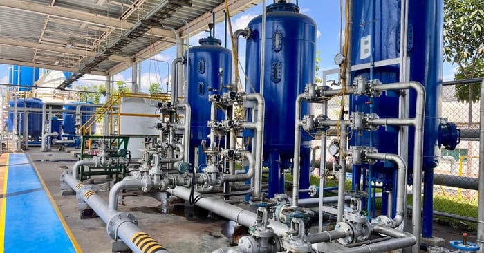 Three large blue industrial air tanks connect to a network of silver pipes, valves, and gauges beneath a metal roof.