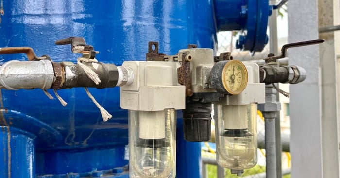 Compressed air filter-regulator units with clear bowls and a pressure gauge connect to piping beside a large blue tank.