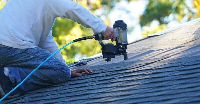 A person in a white shirt and jeans uses a black nail gun with a blue hose to nail shingles to a roof.