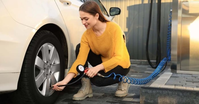 A brown-haired woman in a yellow sweater and black pants uses an air pump with a blue hose to inflate a car tire.