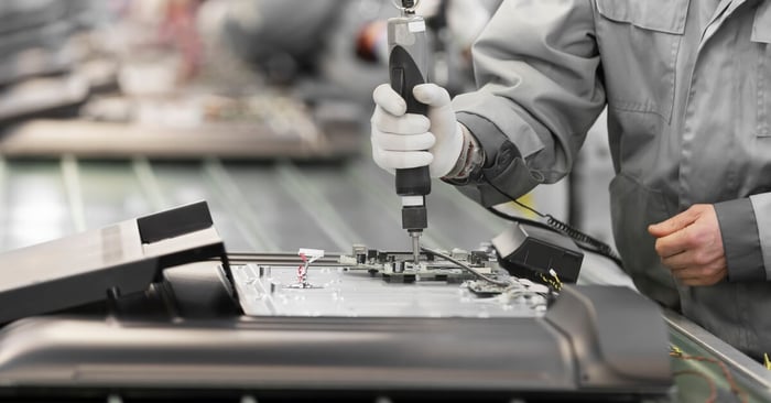 A person in a gray jumpsuit and white gloves assembles electronics with a handheld tool on a manufacturing line.