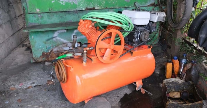 An orange horizontal air compressor with a motor, a gauge, and a green hose sits on a concrete slab.