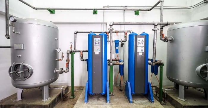 An industrial room with two large silver tanks, blue control units, and a complex network of pipes, gauges, and valves.