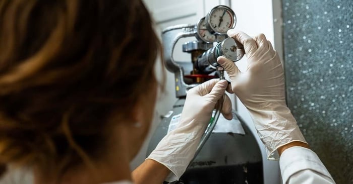 A person in a lab coat adjusts a gas cylinder regulator with gloved hands, featuring pressure gauges and a translucent tube.