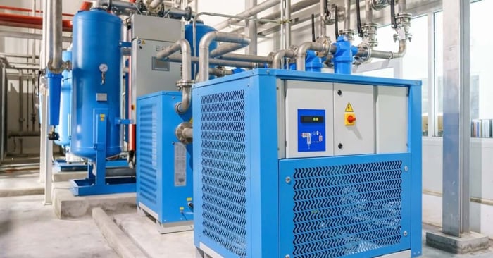 An industrial blue air compressor system with a large tank, control panel, and overhead piping in a factory setting.