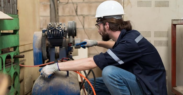 A technician in a hard hat repairing a blue air compressor tank with tools and an orange hose in a workshop setting.