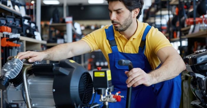 A male technician in blue overalls and a yellow shirt inspects an air compressor motor inside an equipment store.