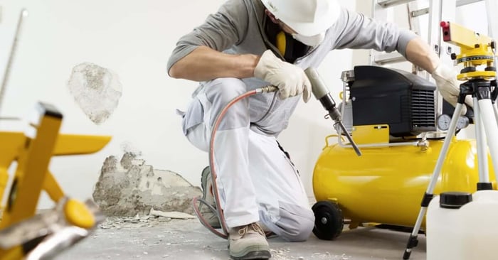 A worker in a hard hat kneels beside a yellow air compressor, using a pneumatic drill on a damaged concrete wall indoors.
