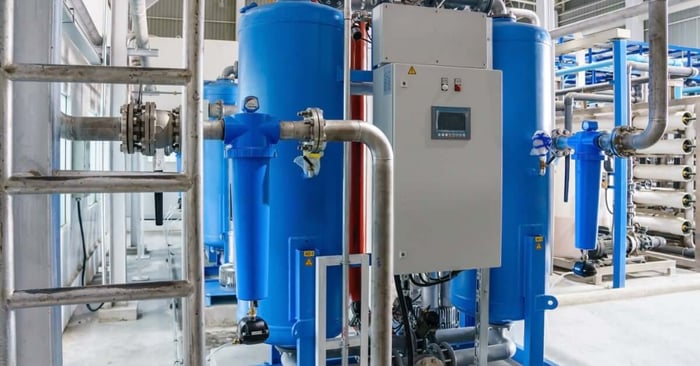 Blue industrial air dryer tanks with a control panel and metal piping system inside a mechanical facility.