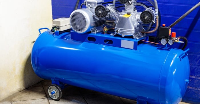 A blue horizontal air compressor tank with a mounted motor and dual-cylinder pump positioned on a workshop floor.