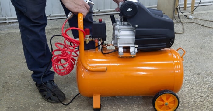 A worker attaching a coiled red hose to a portable orange air compressor with a motor and tank in a workshop setting.