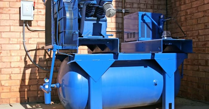 A large blue industrial air compressor mounted beside a brick wall with a storage tank and connected power system.