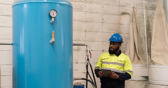 A large blue air receiver tank beside a worker in safety gear inspecting equipment in an industrial facility.