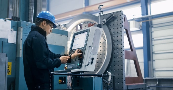 An industrial worker in a blue hard hat operating a CNC machine control panel beside large precision machining equipment.