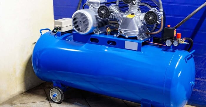 A large blue horizontal air compressor tank with mounted motor, pump assembly, gauges, and wheels on a tile floor.