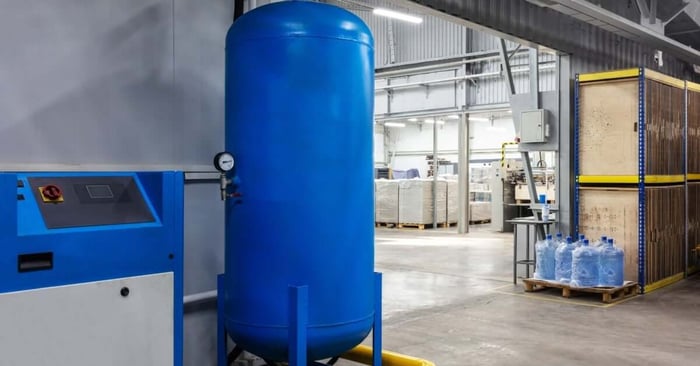 A blue vertical air receiver tank installed beside an industrial air compressor inside a clean manufacturing facility.