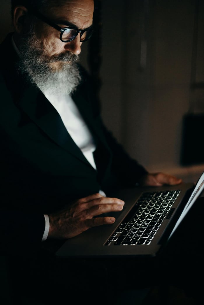 A bearded man works on his laptop in a dark room, illuminated by the screen's glow.