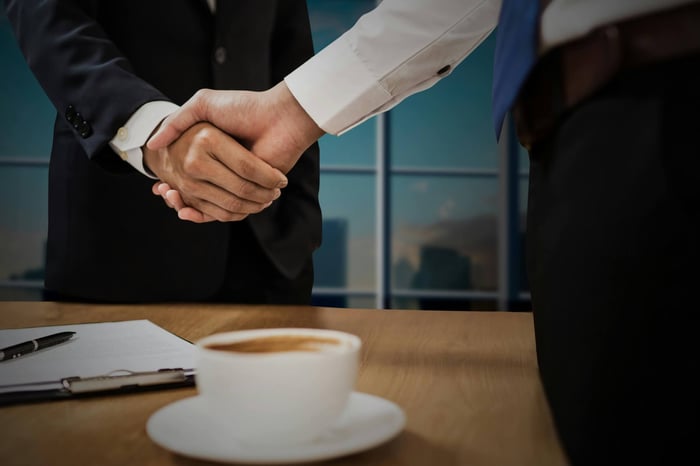 Two professionals shaking hands across a desk with cityscape in the background.